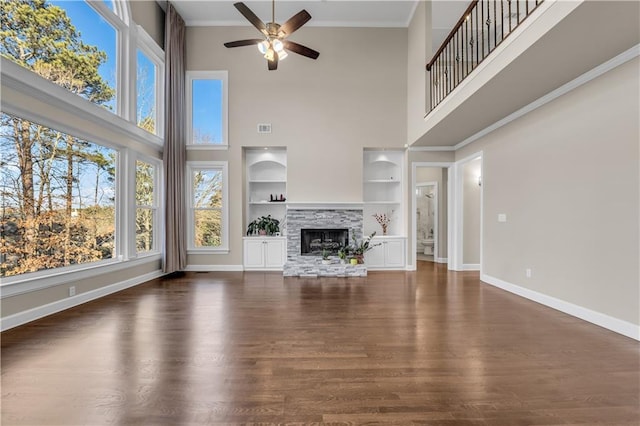 unfurnished living room with a towering ceiling, ceiling fan, built in features, dark hardwood / wood-style flooring, and a stone fireplace