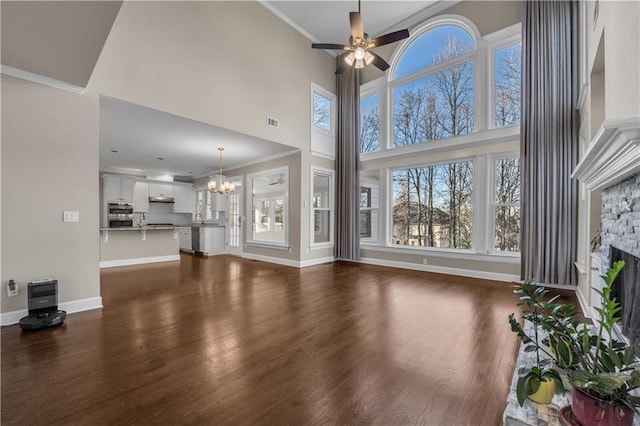 unfurnished living room featuring dark wood-type flooring, a high ceiling, crown molding, ceiling fan with notable chandelier, and a stone fireplace