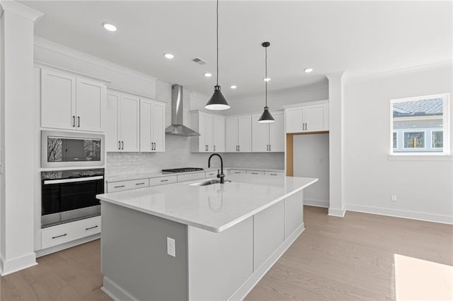 kitchen featuring wall chimney exhaust hood, sink, appliances with stainless steel finishes, an island with sink, and white cabinets