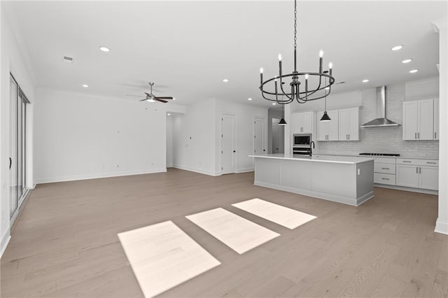 kitchen featuring wall chimney range hood, a kitchen island with sink, hanging light fixtures, stainless steel microwave, and white cabinets