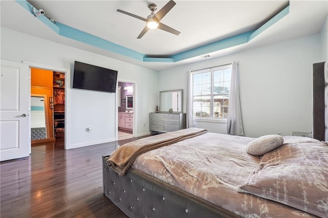 bedroom with a tray ceiling, a walk in closet, a ceiling fan, ensuite bath, and wood finished floors