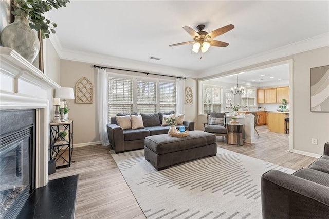 living room with ceiling fan with notable chandelier, a high end fireplace, light wood-style floors, crown molding, and baseboards