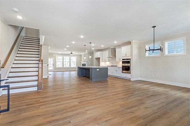 kitchen featuring wall chimney range hood, an island with sink, decorative light fixtures, white cabinets, and ceiling fan with notable chandelier