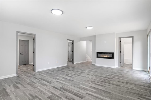 unfurnished living room featuring stairs, a glass covered fireplace, baseboards, and wood finished floors