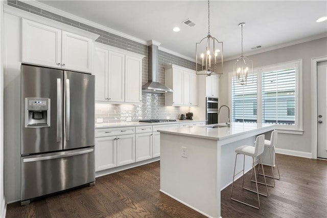 kitchen with a kitchen island with sink, white cabinetry, stainless steel appliances, decorative light fixtures, and wall chimney exhaust hood