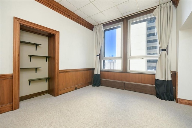 interior space with a drop ceiling, light colored carpet, a wainscoted wall, ornamental molding, and wood walls