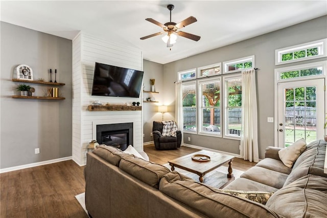 living area featuring a ceiling fan, baseboards, a large fireplace, and dark wood-type flooring