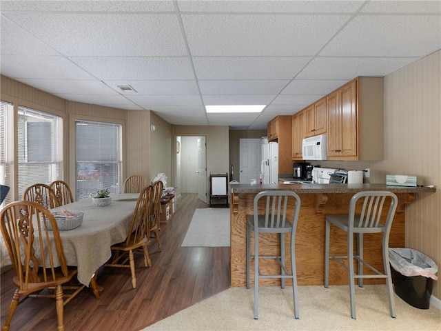 kitchen featuring white appliances, kitchen peninsula, dark hardwood / wood-style flooring, a kitchen bar, and a drop ceiling