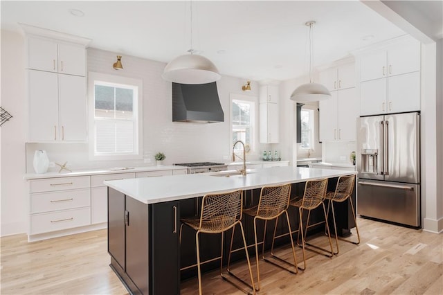 kitchen featuring pendant lighting, an island with sink, white cabinets, high end fridge, and wall chimney exhaust hood