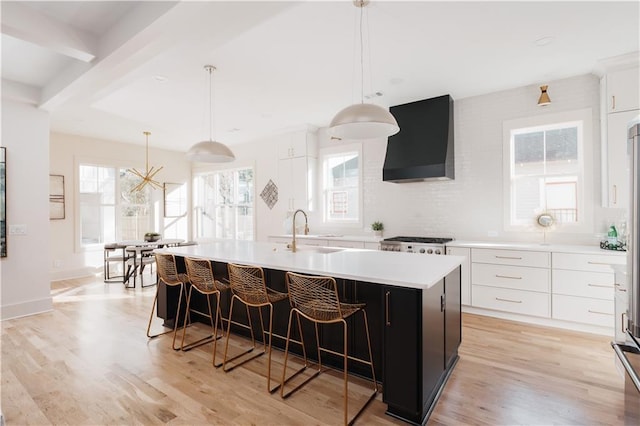 kitchen featuring wall chimney range hood, light hardwood / wood-style flooring, white cabinetry, hanging light fixtures, and an island with sink