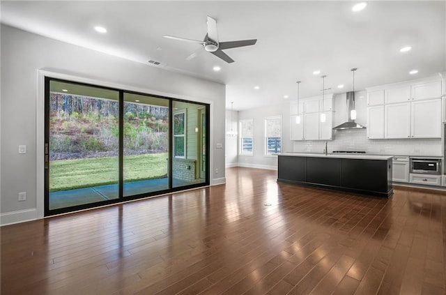 kitchen with hanging light fixtures, wall chimney range hood, a kitchen island with sink, decorative backsplash, and white cabinets