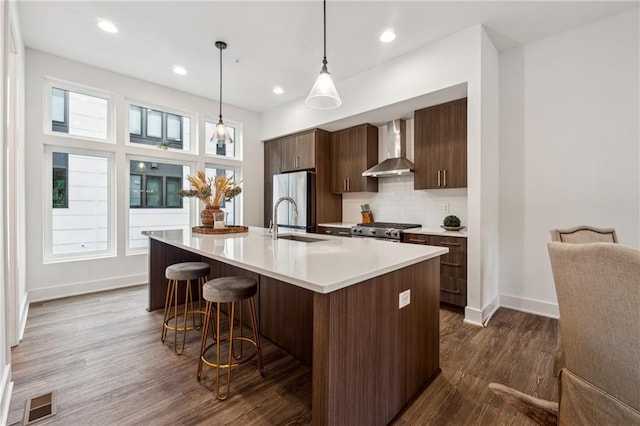 kitchen featuring pendant lighting, a center island with sink, wall chimney range hood, sink, and stainless steel appliances