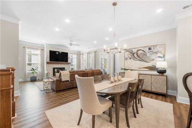 dining area featuring a brick fireplace, ceiling fan with notable chandelier, crown molding, and wood finished floors