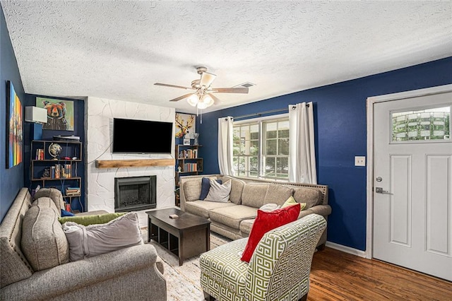 living area featuring wood finished floors, ceiling fan, a stone fireplace, a textured ceiling, and baseboards