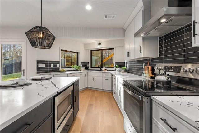 kitchen featuring wall chimney range hood, white cabinetry, stainless steel appliances, light stone countertops, and decorative light fixtures