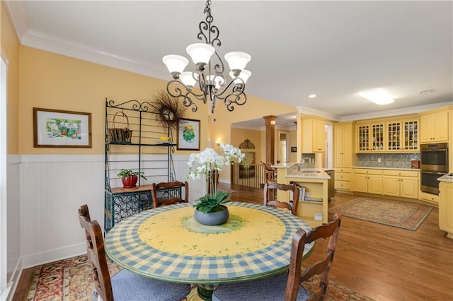 dining room with light wood-style flooring, a wainscoted wall, crown molding, an inviting chandelier, and ornate columns