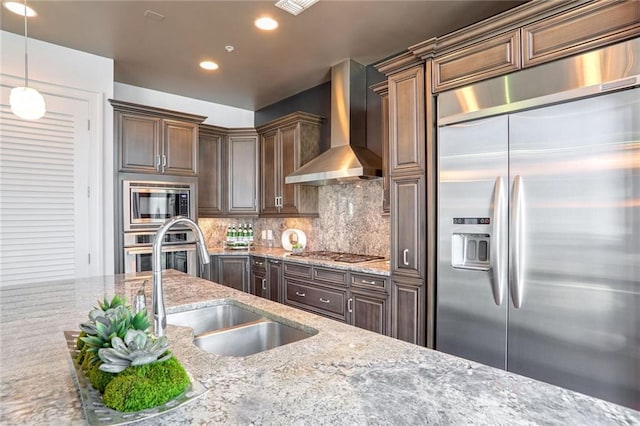 kitchen with pendant lighting, decorative backsplash, built in appliances, light stone counters, and wall chimney exhaust hood