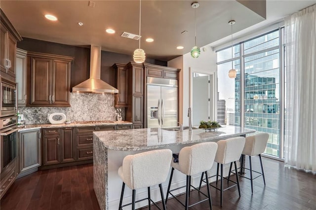 kitchen featuring a kitchen island with sink, wall chimney range hood, decorative light fixtures, and built in appliances