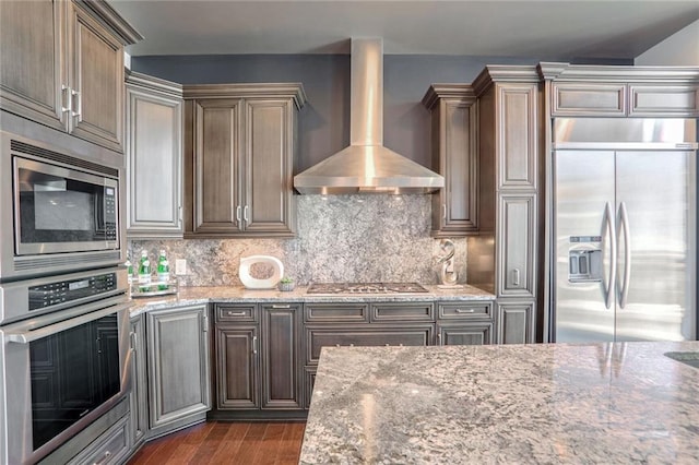 kitchen featuring wall chimney range hood, backsplash, built in appliances, light stone counters, and dark hardwood / wood-style flooring