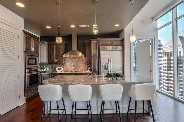 kitchen with wall chimney exhaust hood, built in appliances, pendant lighting, a kitchen island with sink, and backsplash