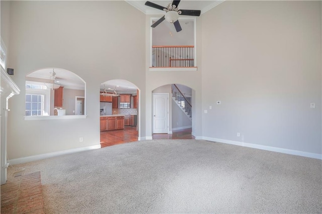 unfurnished living room with ceiling fan, light colored carpet, ornamental molding, and a high ceiling