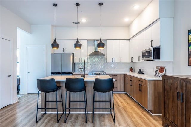kitchen featuring white cabinets, light wood-style floors, wall chimney exhaust hood, stainless steel appliances, and a kitchen bar