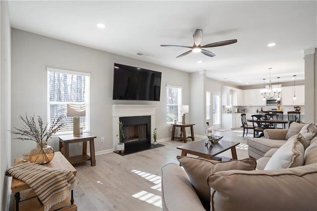 living room featuring a fireplace with raised hearth, baseboards, light wood-type flooring, recessed lighting, and ceiling fan with notable chandelier