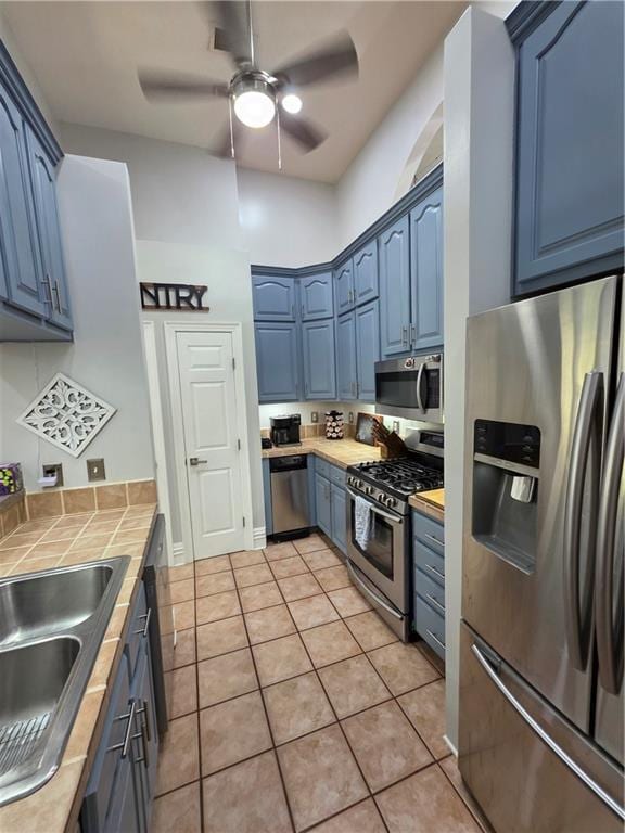 kitchen with blue cabinets, stainless steel appliances, light tile patterned floors, ceiling fan, and tile countertops