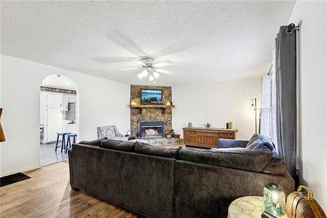 living room featuring ceiling fan, a stone fireplace, a textured ceiling, and light hardwood / wood-style floors