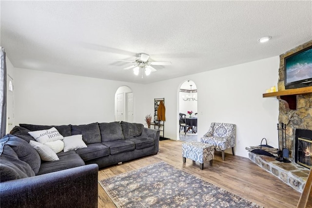 living room featuring ceiling fan, a stone fireplace, a textured ceiling, and light hardwood / wood-style flooring