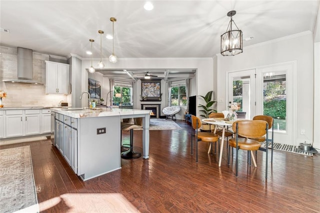 kitchen featuring a kitchen island with sink, wall chimney range hood, pendant lighting, white cabinetry, and dark hardwood / wood-style flooring