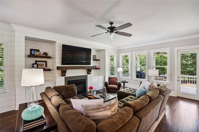 living area featuring baseboards, a glass covered fireplace, ceiling fan, dark wood-style flooring, and crown molding