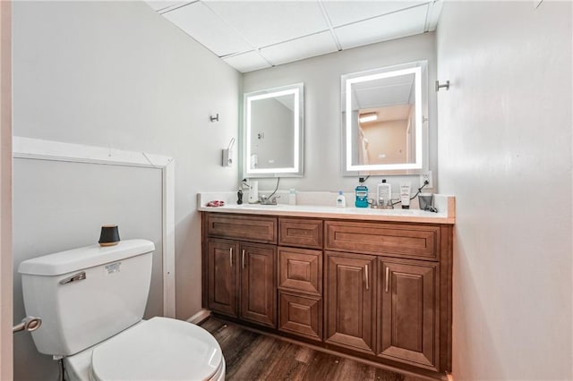bathroom with double vanity, wood finished floors, baseboards, toilet, and a drop ceiling