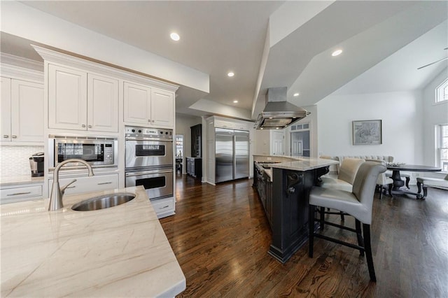 kitchen featuring light stone countertops, a sink, built in appliances, white cabinetry, and island range hood