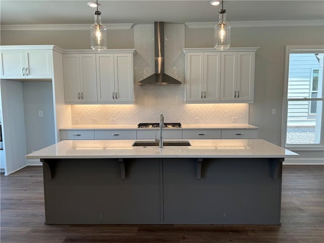 kitchen featuring decorative light fixtures, white cabinetry, wall chimney exhaust hood, and an island with sink