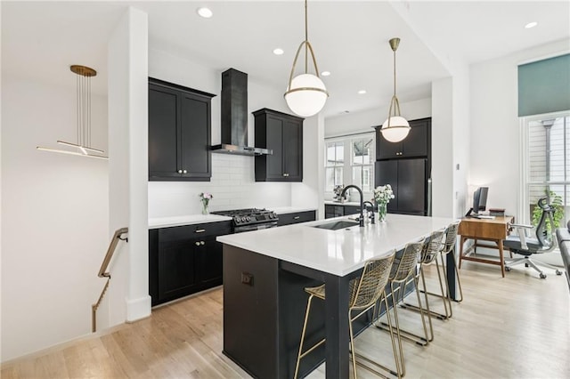 kitchen with wall chimney exhaust hood, dark cabinets, light countertops, light wood-style flooring, and a center island with sink