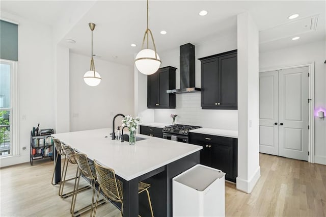 kitchen with wall chimney range hood, dark cabinets, stainless steel range with gas stovetop, a center island with sink, and recessed lighting