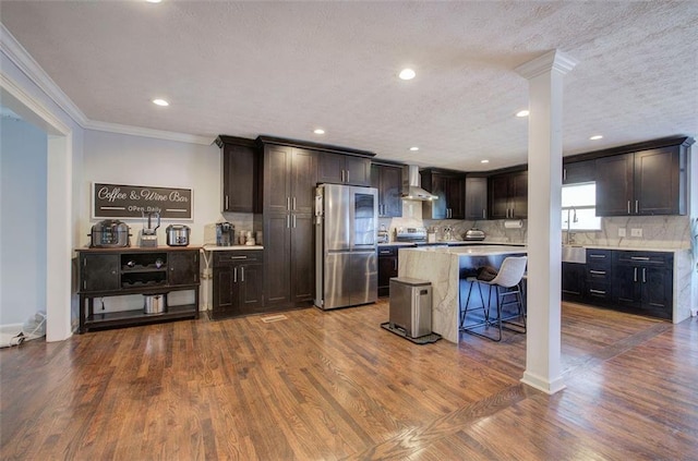 kitchen featuring a breakfast bar area, stainless steel appliances, light countertops, a kitchen island, and wall chimney range hood