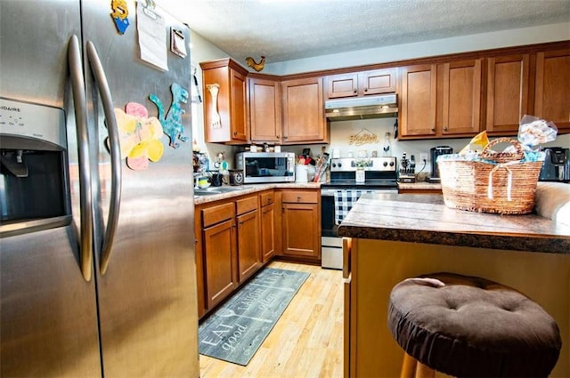 kitchen with a textured ceiling, under cabinet range hood, stainless steel appliances, light wood finished floors, and brown cabinetry