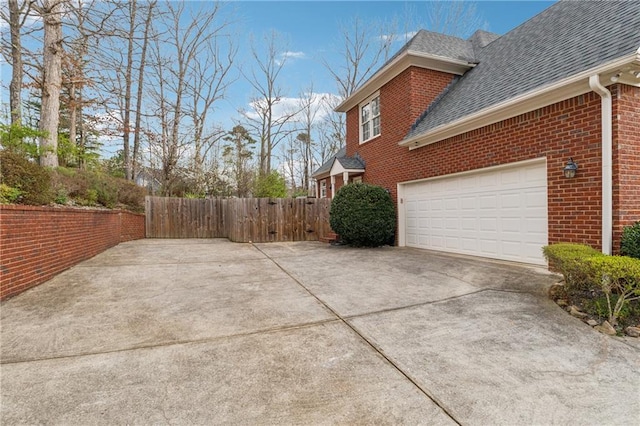 view of side of property featuring fence, concrete driveway, a shingled roof, a garage, and brick siding