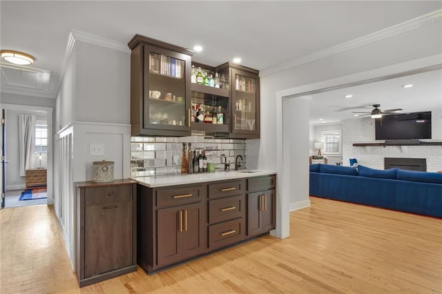 indoor wet bar with light wood-type flooring, ceiling fan, tasteful backsplash, a brick fireplace, and ornamental molding