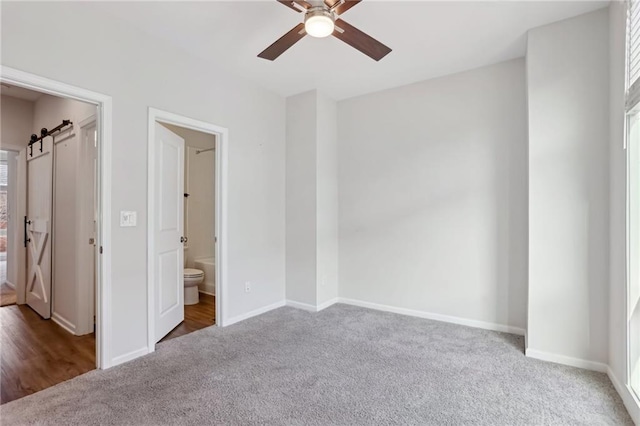 unfurnished bedroom featuring ceiling fan, ensuite bathroom, a barn door, and carpet flooring