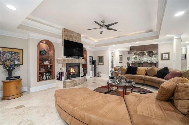 living room featuring a tray ceiling, a fireplace, ornamental molding, ceiling fan, and baseboards