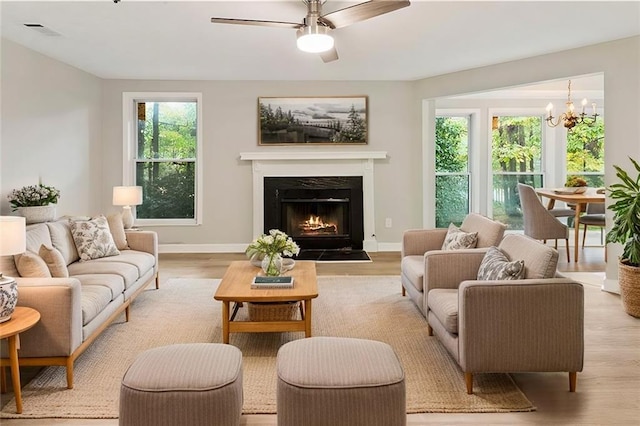 living area with a warm lit fireplace, light wood-style flooring, ceiling fan with notable chandelier, visible vents, and baseboards