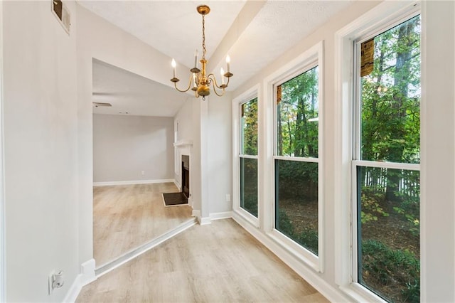 unfurnished dining area featuring a fireplace with flush hearth, visible vents, baseboards, light wood-type flooring, and an inviting chandelier