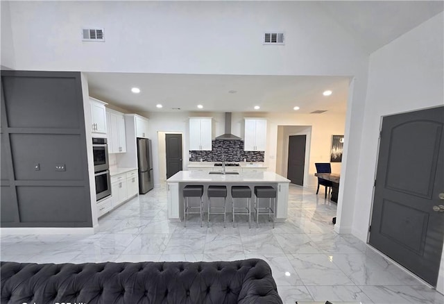 kitchen with wall chimney exhaust hood, a kitchen breakfast bar, stainless steel appliances, a kitchen island with sink, and white cabinets