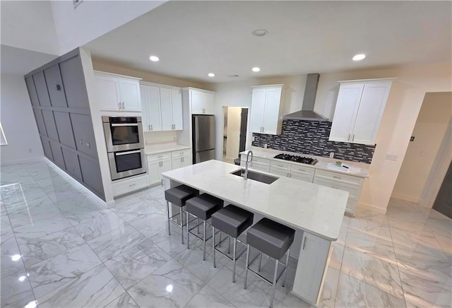 kitchen with sink, a breakfast bar area, a center island with sink, appliances with stainless steel finishes, and wall chimney range hood