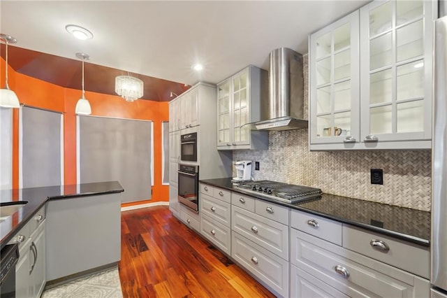 kitchen with hanging light fixtures, backsplash, wall chimney exhaust hood, dark countertops, and glass insert cabinets