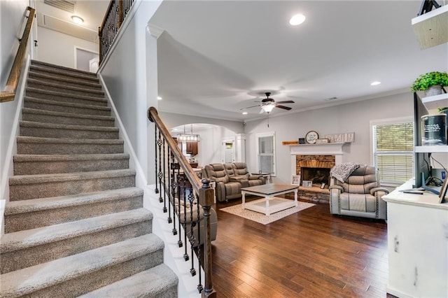 living area with arched walkways, a ceiling fan, stairs, a stone fireplace, and dark wood finished floors