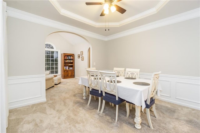 dining area with arched walkways, a raised ceiling, light colored carpet, a wainscoted wall, and ornamental molding